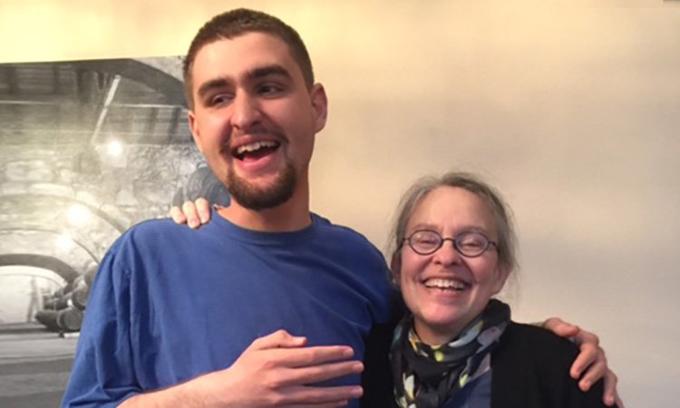 Danny & Nina A young man with short brown hair and a small beard stands with his arm around a woman, his mother, while both of them laugh. He is a full head taller than her.