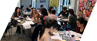Groups of people gathered around several tables. Some groups are speaking to each other. Others are working on papers in front of them.