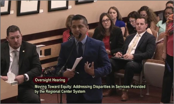Abner A Latino man speaks at a microphone, wearing a suit and holding papers. People sit in rows behind him. Text on image reads: Oversight Hearing. Moving Toward Equity: Addressing Disparities in Services Provided by the Regional Center System
