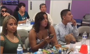 Jared Two Latina women and a Latino man with a disability sit at a table with food, looking off toward a presentation